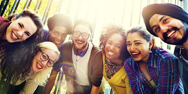 Seven young adults of different ethnicities stand smiling with their heads together, trees and sunshine in the background