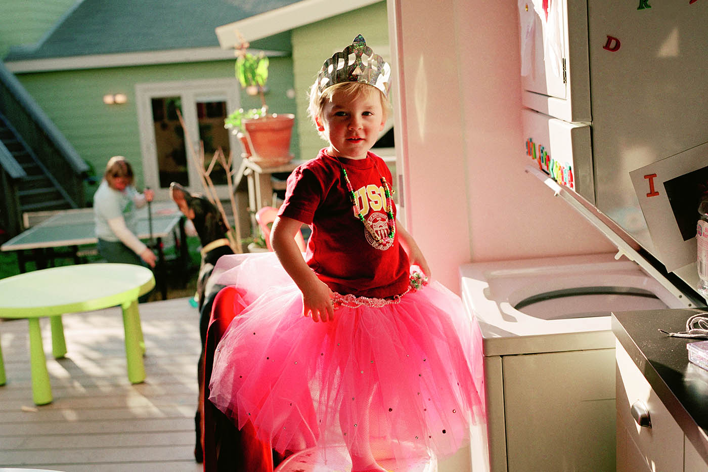 A young boy wearing a pink tutu and a silver tiara is standing on a kitchen chair in front of an open washing machine. The door is open behind him and a woman can be seen sweeping outside.