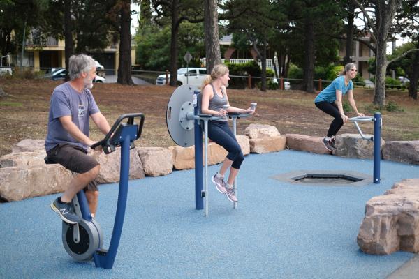 One man and two women using exercise equipment at park with cars and houses in the background