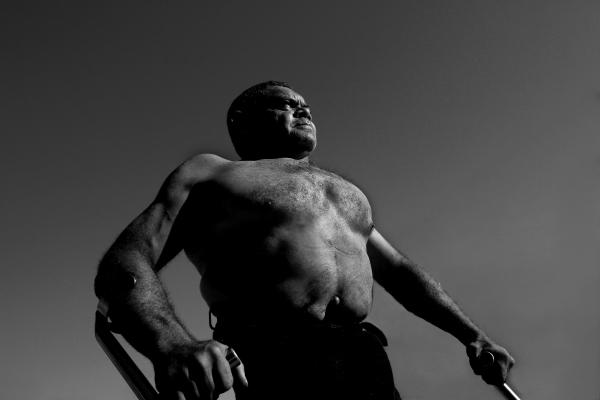 Black and white image of a First Nations man. He stands shirtless against an empty sky, holding crutches and looking into the distance.
