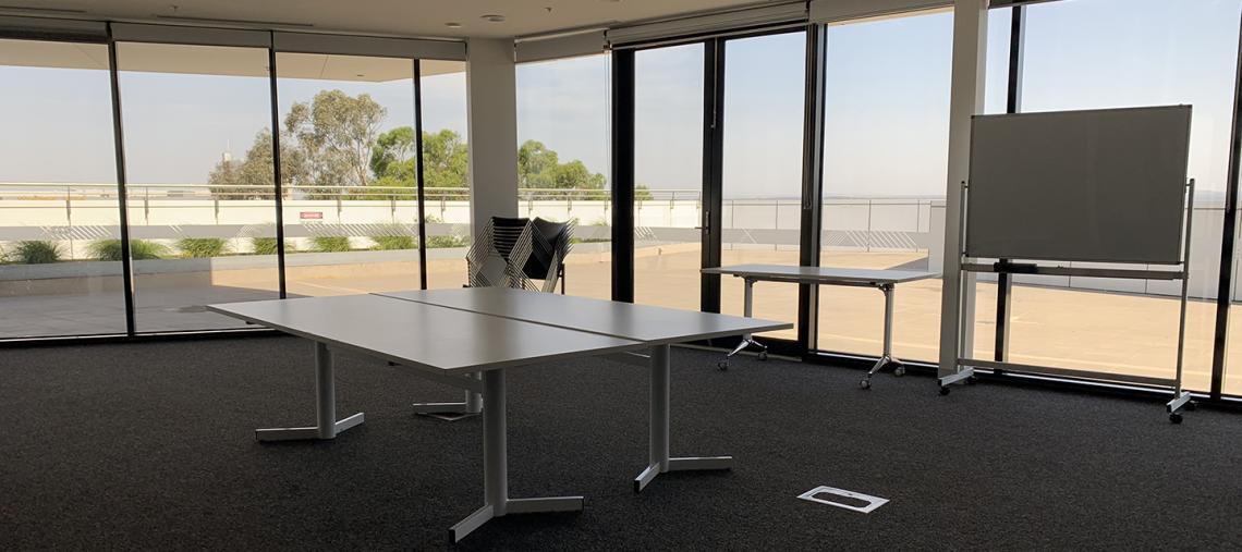 Interior shot of an empty meeting room with full sized windows and glass doors, containing three tables, a stack of chairs and a white board.