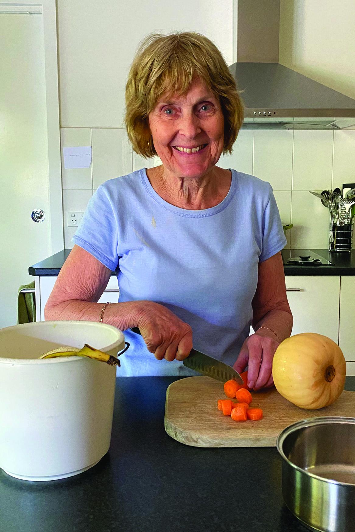 An older woman wearing a blue tshirt stands smiling at a kitchen bench while chopping vegetables. A light brown FOGO caddy is on the bench.