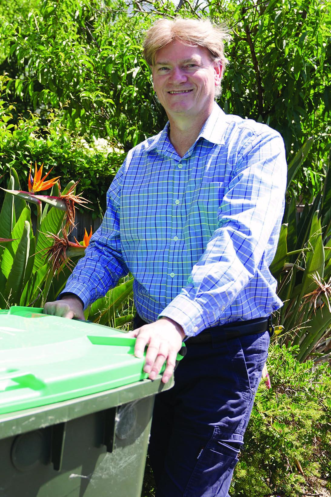 A smiling middle-aged man wearing a blue checked shirt pushes a green waste bin in a garden setting