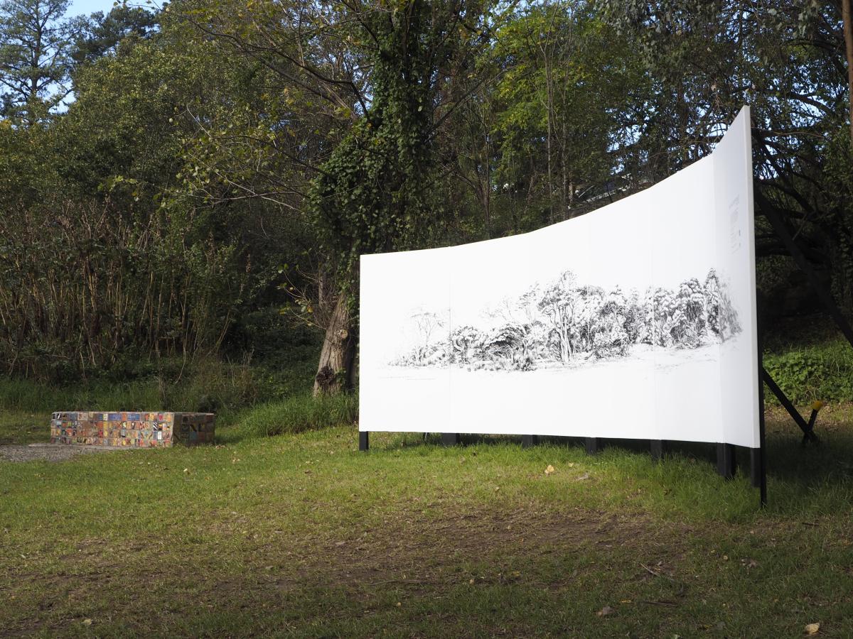 Photograph of a curved white billboard featuring a detailed black landscape drawing installed in parkland