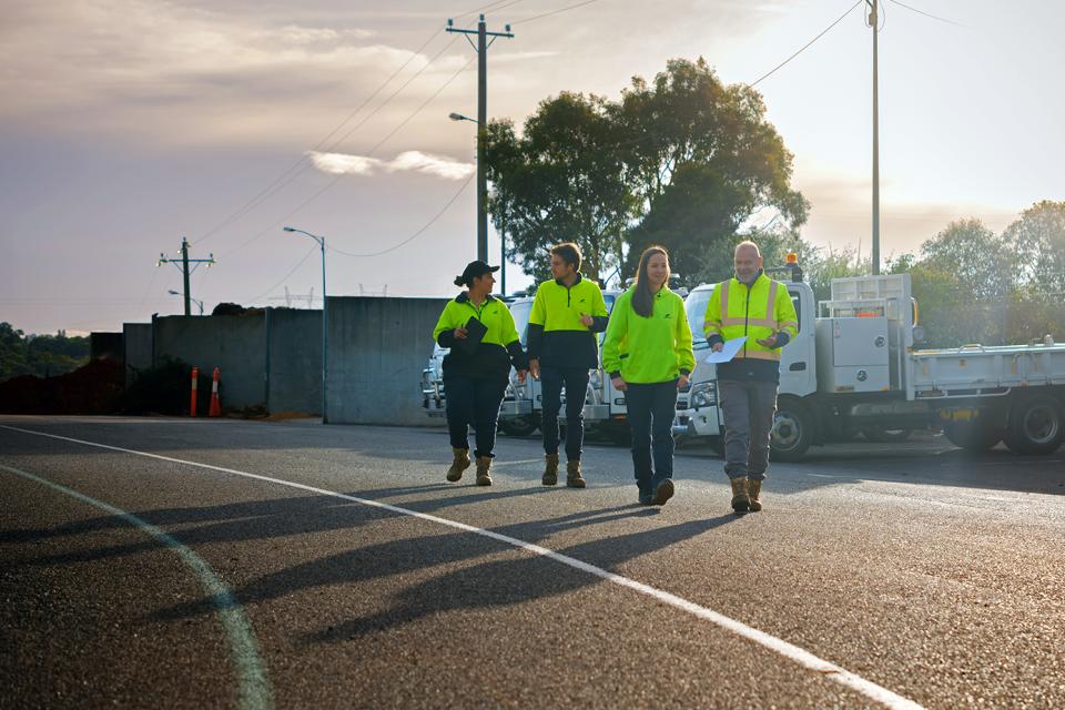 Four people in yellow safety clothing and work boots walk down an empty road talking to each other. Trucks can be seen in the background.