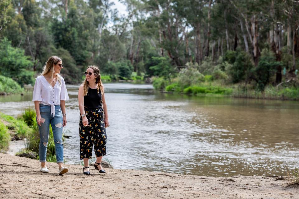 Two young women walk beside a wide river surrounded by gum trees and shrubs. They are looking at each other and smiling.