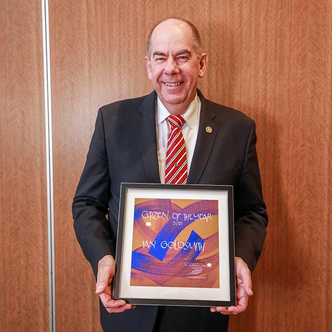 Man wearing a suit stands smiling and holding a framed award in front of his body