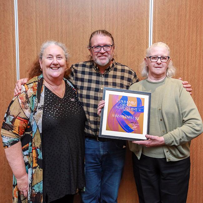 Two women and one man stand smiling and holding a framed award in front of their bodies