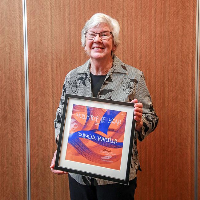Older woman with glasses, wearing a brown and black shirt stands smiling and holding a framed award in front of her body