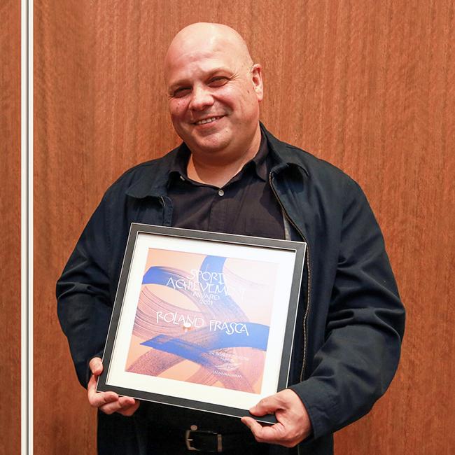 Man wearing a black shirt stands smiling and holding a framed award in front of his body