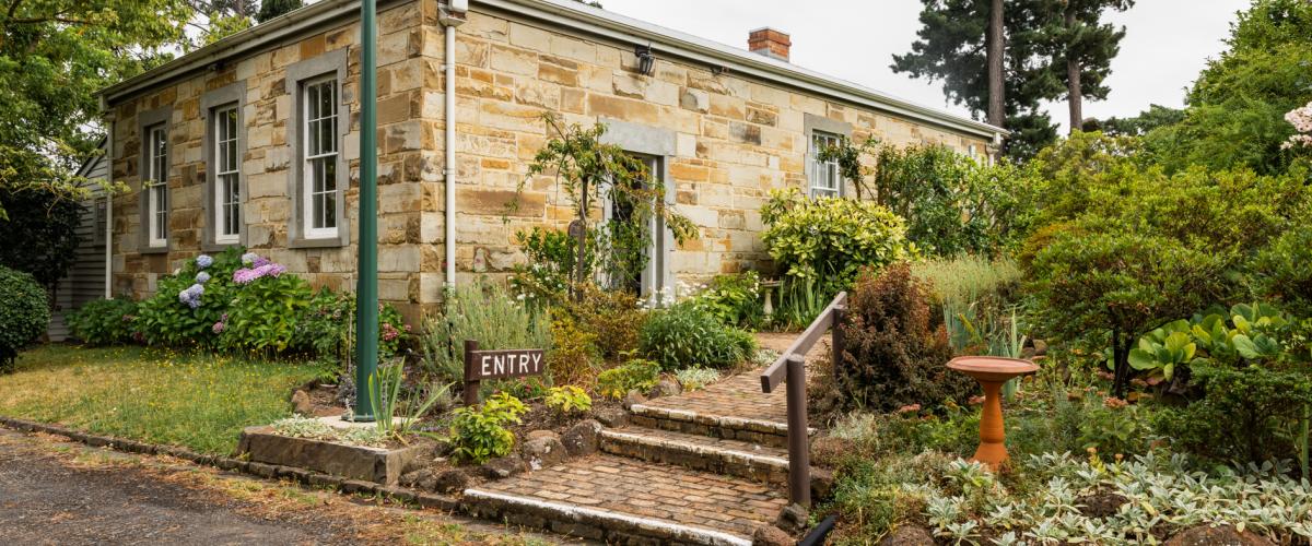 A restored sandstone building in a cottage garden setting. A small sign with the word 'entry' sits at the base of a path leading to the side door of the building.