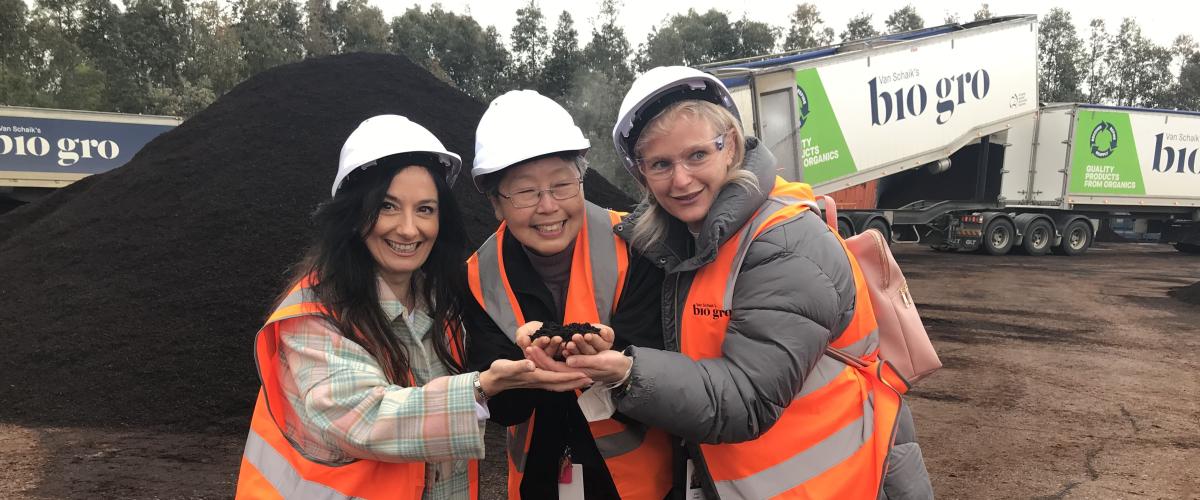 Three Manningham councillors smiling and wearing hard hats, stand holding a handful of compost. In the background is a large pile of compost and a Bio Gro truck.