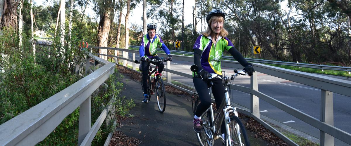A man and woman wearing helmuts and hi vis jackets cycling across a bridge whilst facing the camera