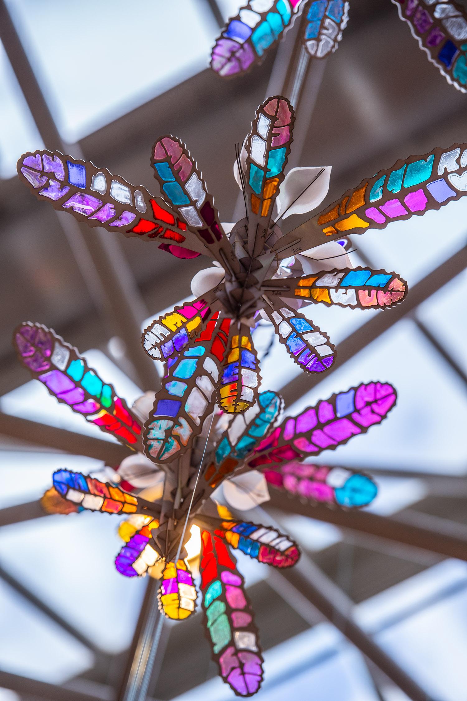Small handmade flower sculpture handing from a glass ceiling. The sculpture is made from a cardboard frame and the petals are made from various coloured material. The light shines through the petals and illuminates the colours. 