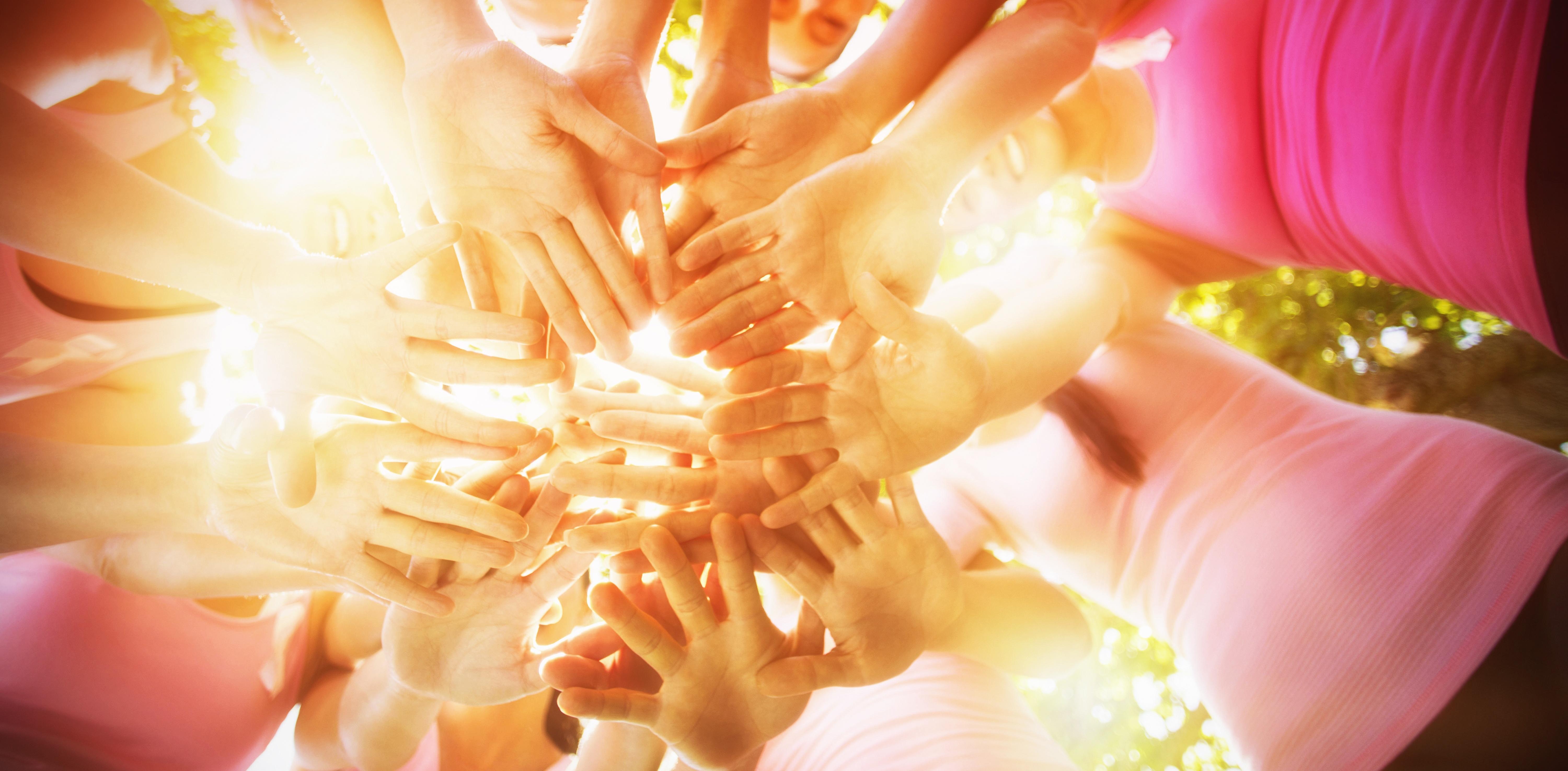 Many women's hands placed together in a circle, illuminated by sunlight shining above them