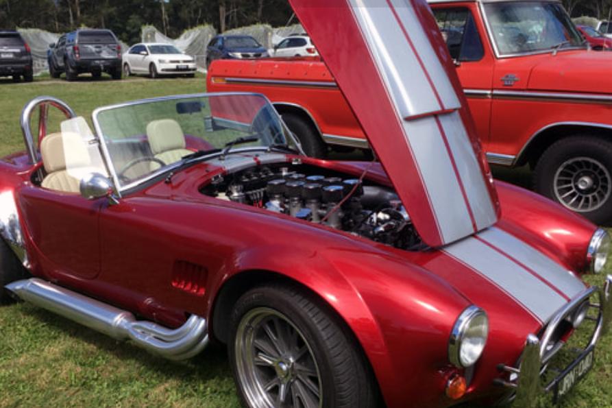 classic red sportscar with white stripe, bonnet is raised showing engine, parked on a green field and surrounded by other cars