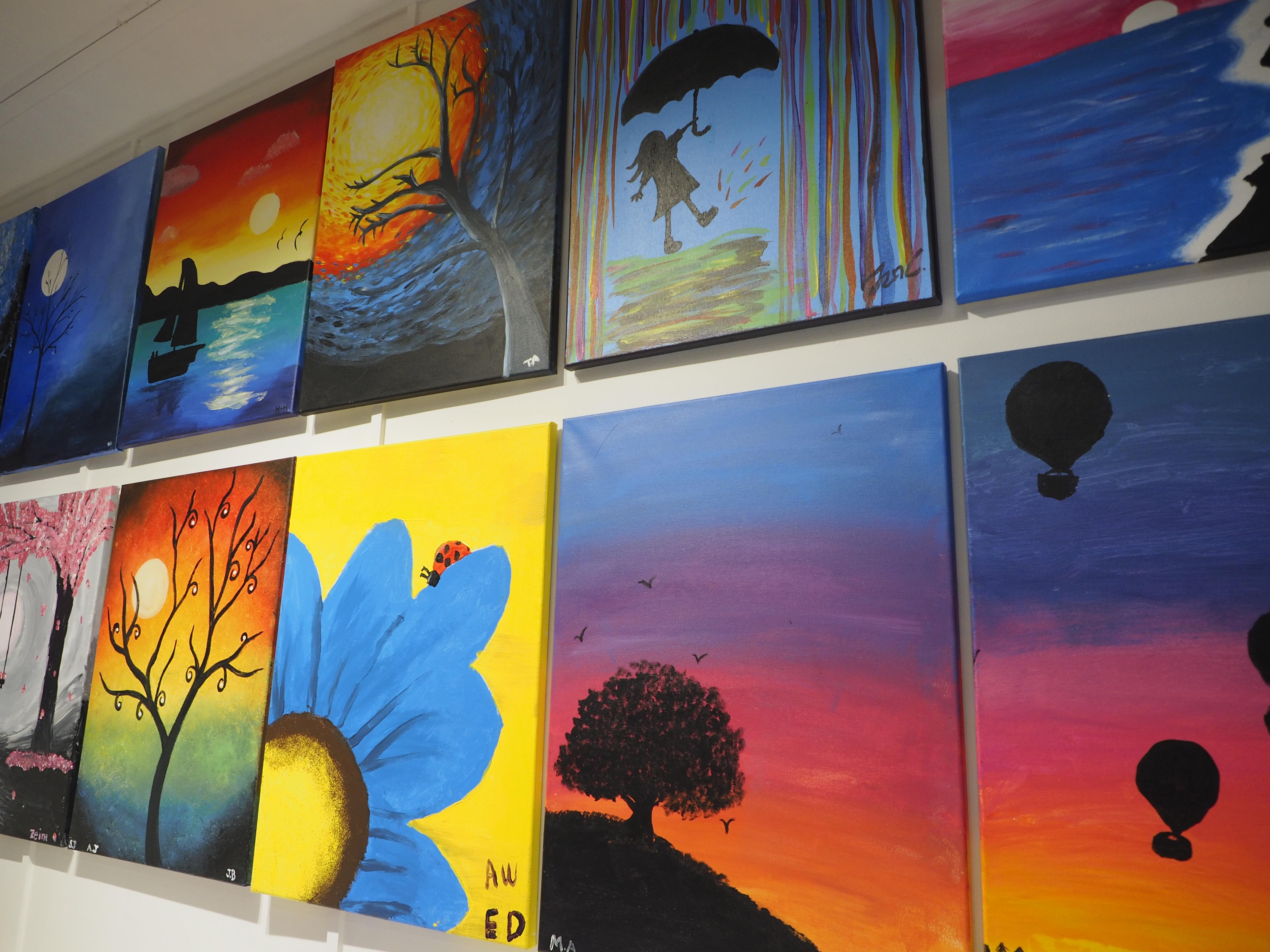 Photograph of several small, brightly coloured paintings depicting images like trees and seascapes hung tightly next to one another on a wall