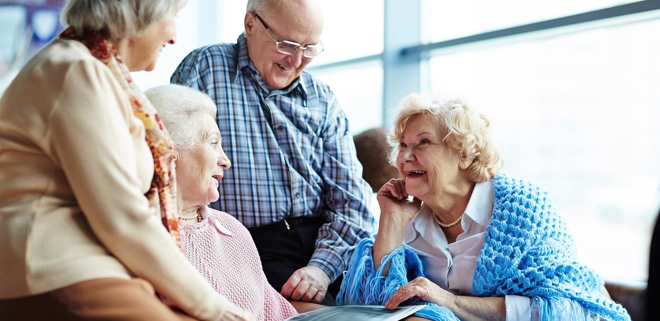 4 elderly people in conversation around armchair laughing