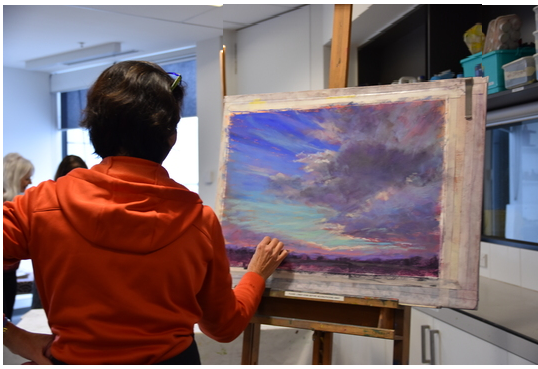 Photograph of a person in a red to drawing a landscape of clouds with a low horizon line in pastels