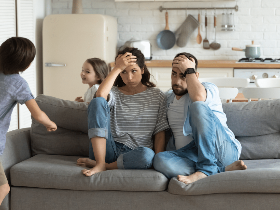 Stressed man and woman sitting on sofa with kids running around