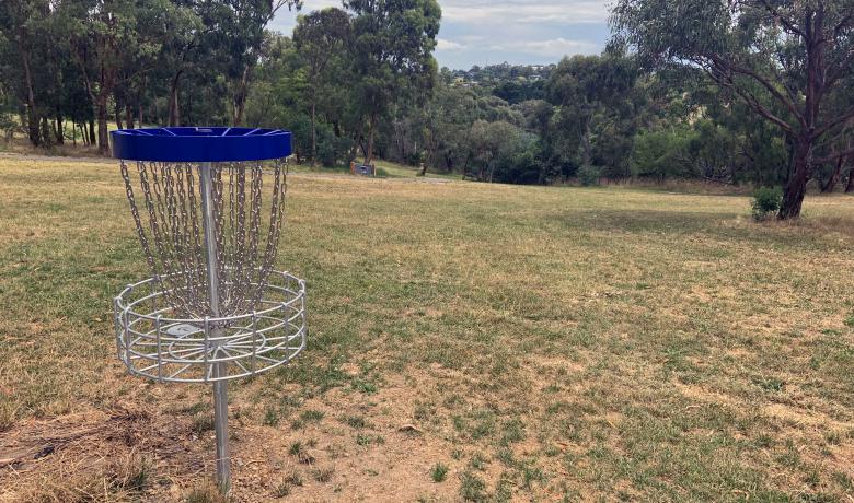 Metal disc golf catcher basket with chains sits at the top of a grassy hill with native trees to the sides and in the distance