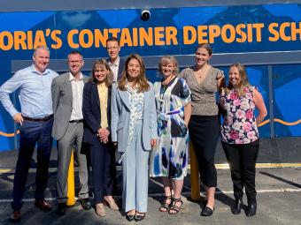 A group of eight smiling people dressed in corporate clothing stand in a carpark in front of a blue sign with orange writing that reads 'Victoria's container deposit scheme'.