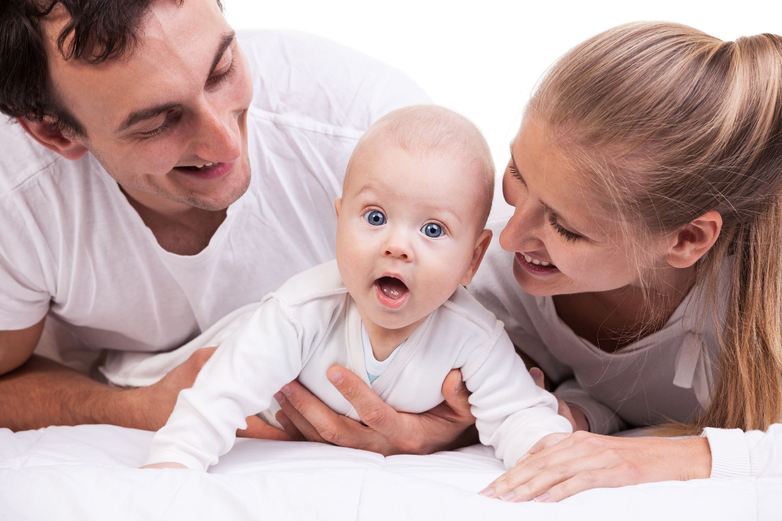 two smiling adults look down at a baby lifting itself up on its arms